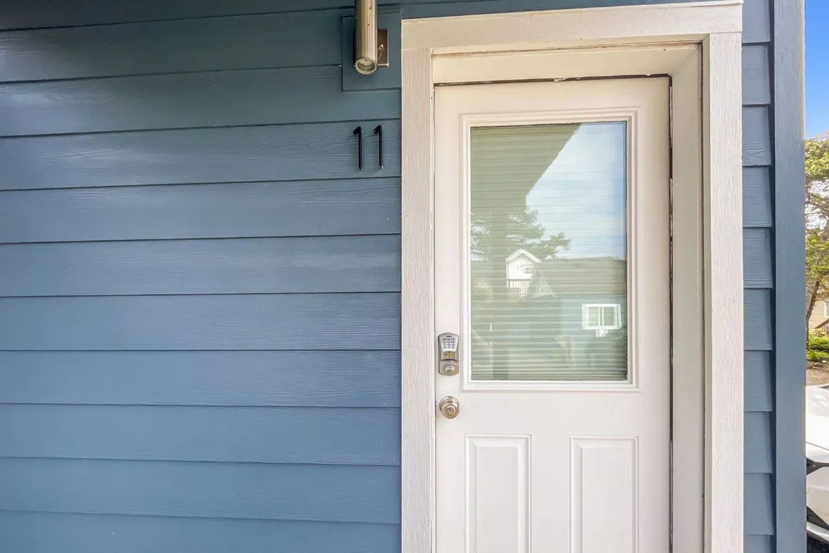 The image shows the exterior entrance of a unit at The Ocean Rogue Inn, with a clearly numbered door (number 11) and blue siding. The scene is bright and welcoming, featuring the entryway and a portion of the outdoor space connected to the unit.