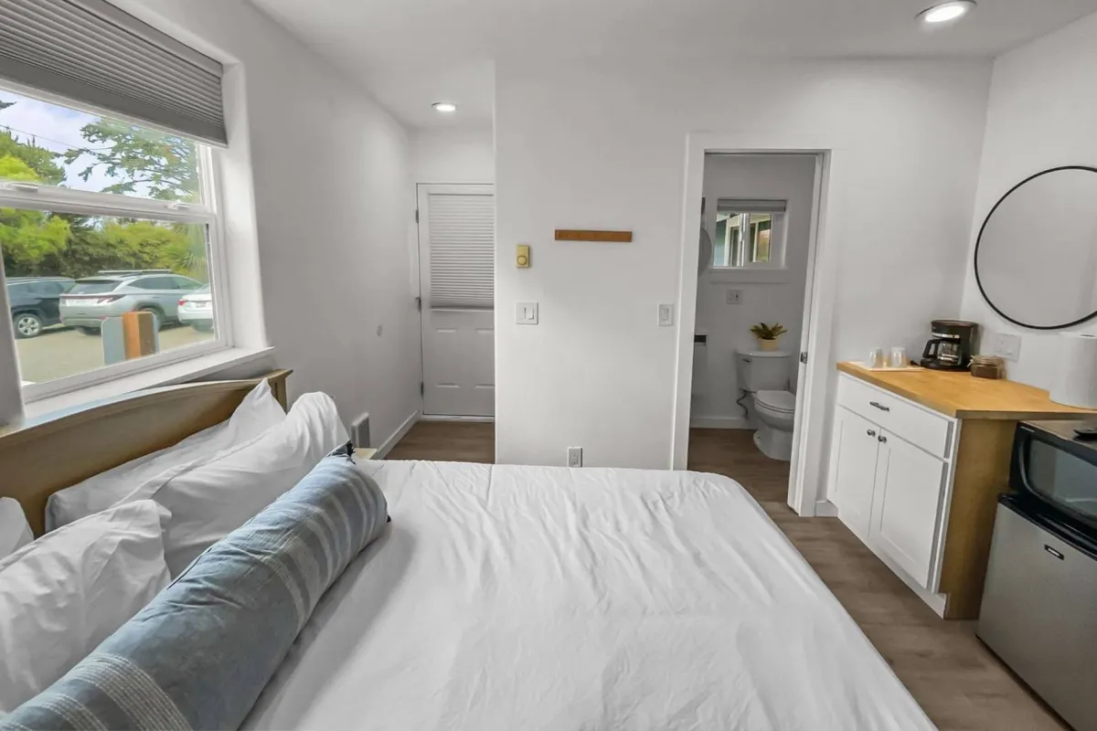 The image showcases a bright and inviting interior of a one-bedroom condo. The bed is neatly made with plush pillows, positioned to catch the natural light streaming in from a window that offers glimpses of the Oregon coast. A compact kitchenette is visible, ideal for preparing small meals, and there's an open door leading to the bathroom. The overall atmosphere is cozy and restful, highlighting the comfort and intimacy of the space.