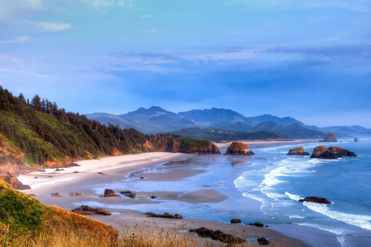 An inviting outdoor scene featuring a sandy beach backed by rocky coastline and distant mountains. As the sun sets, the glow illuminates the shoreline, and a bonfire area is visible, suggesting a space for guests to gather, socialize, and enjoy the oceanfront ambiance together.