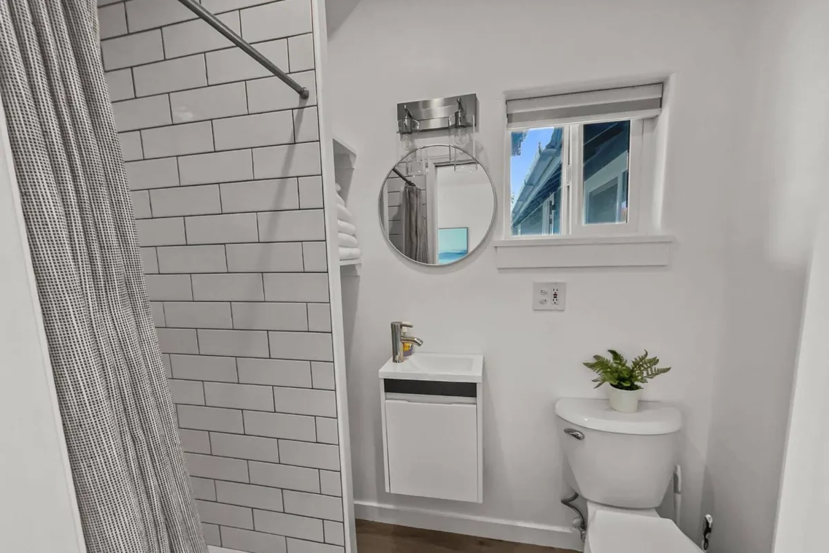 This image features a modern, serene bathroom with a round mirror, a sleek sink, a walk-in shower, and natural light entering through a window. The space is bright and clean, reinforcing the idea of a newly renovated, private bathroom.