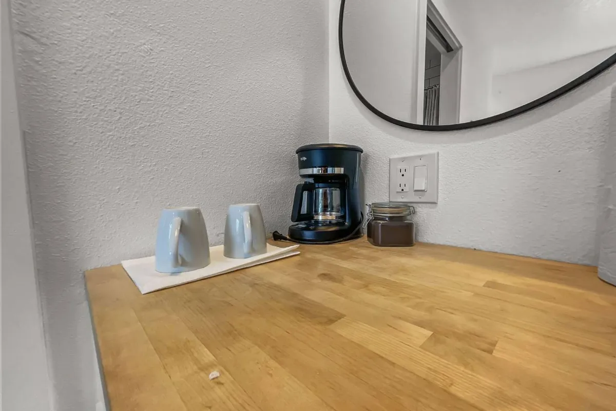 The image features a cozy kitchen corner with a coffee maker, blue cups neatly arranged on a wooden countertop, and a mirror reflecting warm natural light. The scene evokes the pleasure of brewing a fresh cup of coffee, perhaps before stepping out to explore the seaside or after returning from a romantic stroll.