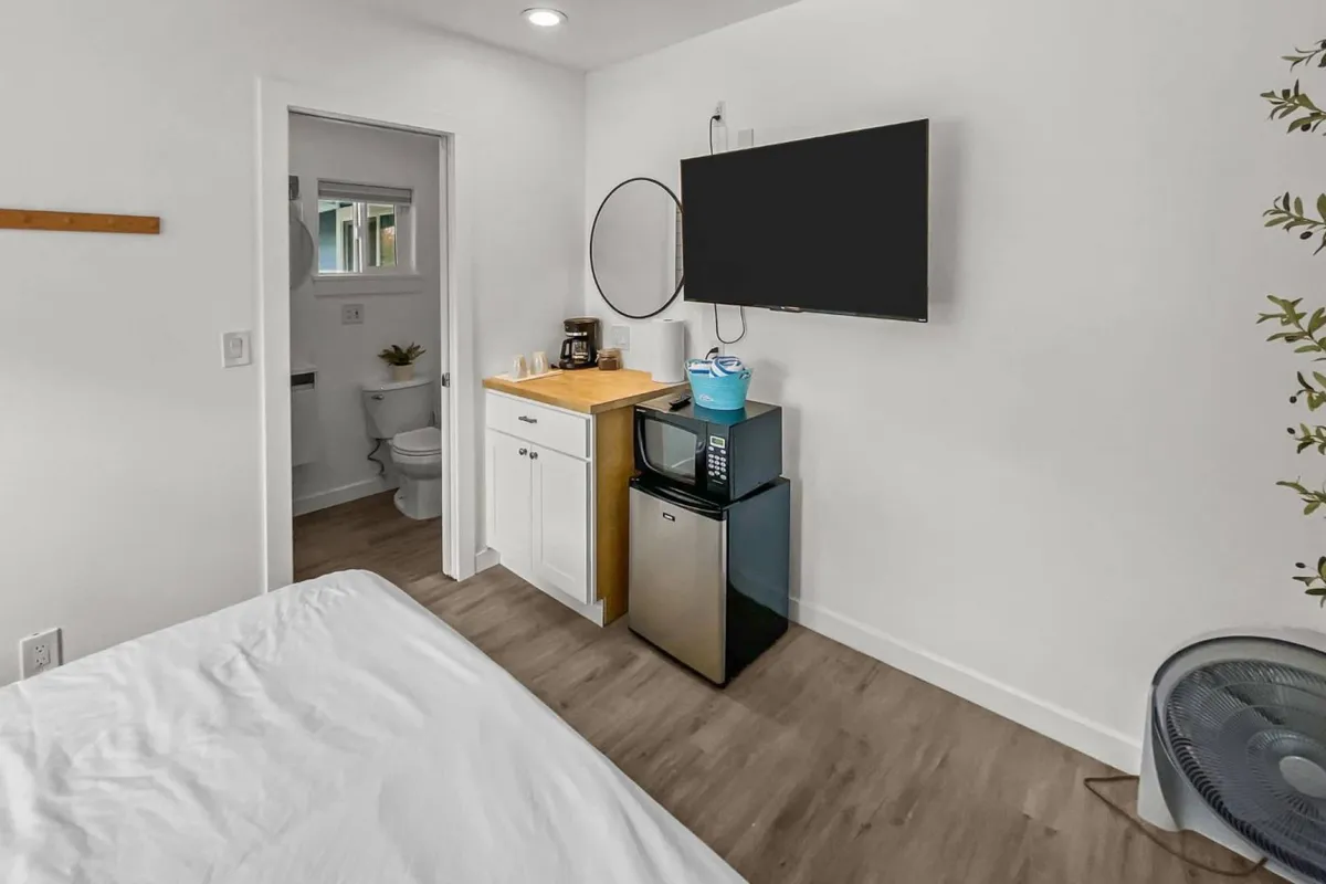 This image shows a comfortable king bed with crisp white linens, a flat-screen TV mounted on the wall, a mini-fridge, and a coffee station. The color palette is calming with soft coastal hues and modern furnishings. The space feels open and inviting, ideal for relaxation and unwinding after a day on the beach.